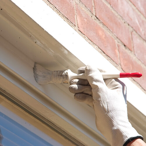 Person's hand wearing glove painting white with paint brush on white framing under red brick wall