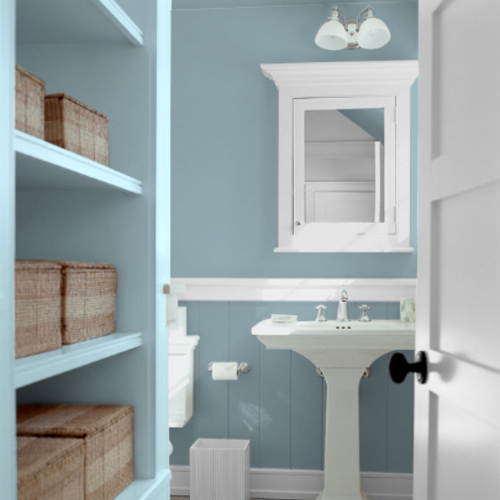 Bathroom with light blue walls and shelves with white sink, framing and mirror with white door on right and brown boxes on left on shelves