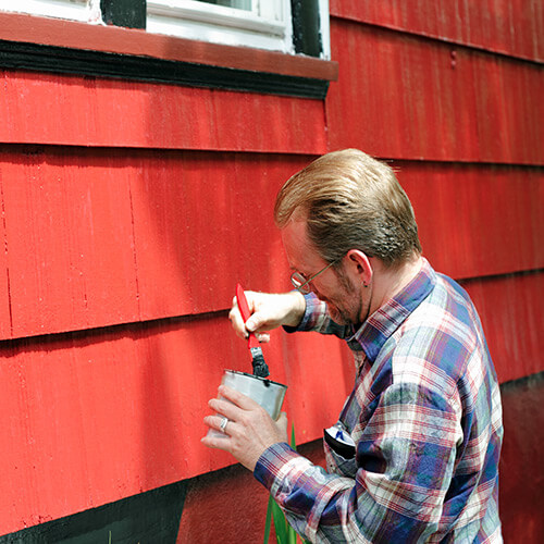 Man with paint bucket dipping paint brush to paint red onto paneled wall