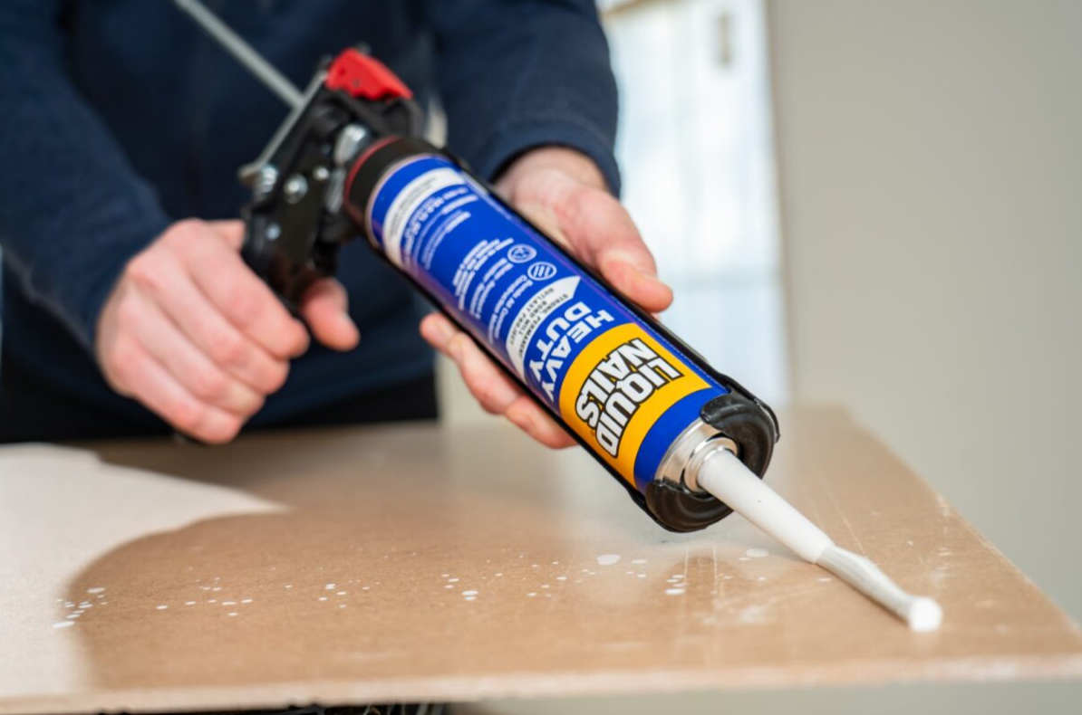 Person holding Liquid Nails adhesive tube applying it on wooden table