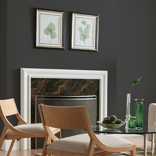 Room with black wall with fireplace and paintings hanging above, and brown wooden chairs 
