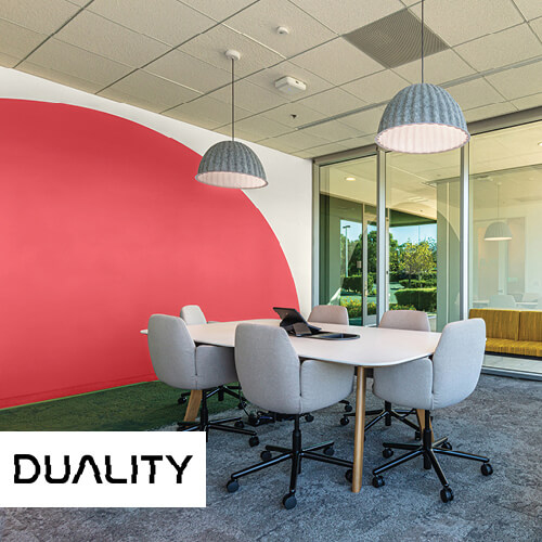 Room with white table, gray chairs, floors, and hanging lights, tan ceiling, red and tan wall with green carpet and large windows to another room with "Duality" on bottom left