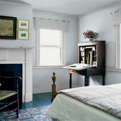 Bedroom with white walls and blue panel floor with fireplace and wooden desk with bed with green blanket, bright windows, and paintings