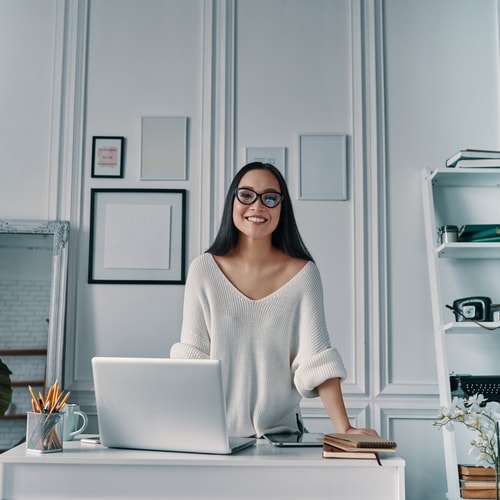 Woman with black hair and glasses wearing white shirt standing and smiling at camera in all white room with paintings, shelves, desk, laptop, and other small items