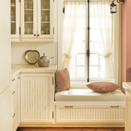 Cream color kitchen with steel teapot and pan on counter with dull salmon color pillows and wall on left with large, curtained window