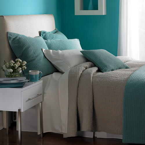 Zoom of top of bed with gray and cyan colors, cyan walls, painting on wall, white side table with small items and window shining on everything from right
