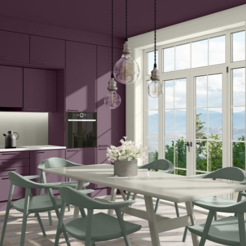Room with dark purple walls and long white table and light gray chairs with white framing on glass back door, with hanging lights and white potted flowers in center