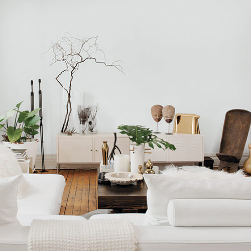 Livingroom with white couches, white wall, and white side tables with brown wooden flooring and coffee table, with potted plants and wooden decoratives in background and around