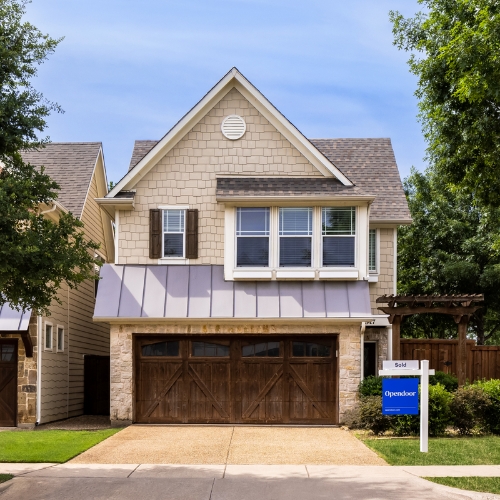 Light brown house with white and dark brown accents and pillars with brown garage door, white sign post with blue Opendoor sales sign, with fence, bushes, and trees around house