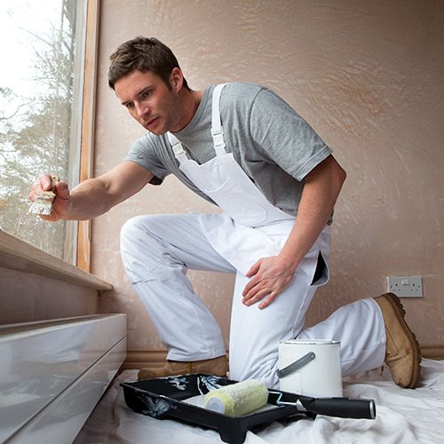 Dark-haired man with gray shirt, white overalls, and tan boots kneeling on ground painting with small paint brush with roller and paint can on ground tan walled room with window 