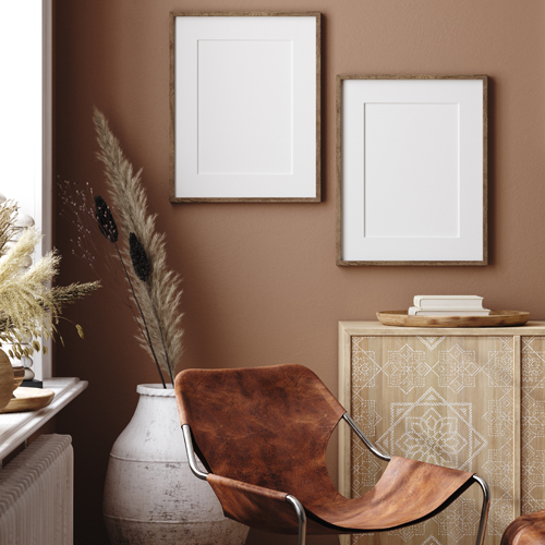 Section of room with brown wall, white paintings hung up, tan table with books on top, white potted plant, brown chair and windowsill on left
