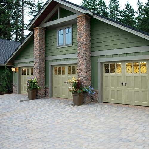 Dark green paneled house with light green garage doors with stone rock pillars with potted plants and stone brick driveway
