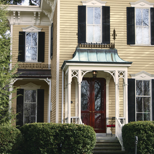Yellow house with windows with black shutters, green mini overhead structure over dark brown front door, with steps and large bushes at bottom
