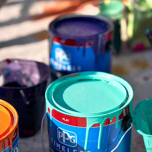 Open PPG paint cans all dripping paint on sides with bright cyan at the foreground, orange on the left, and purple in the back with a blurred background of other paints, all on a concrete ground